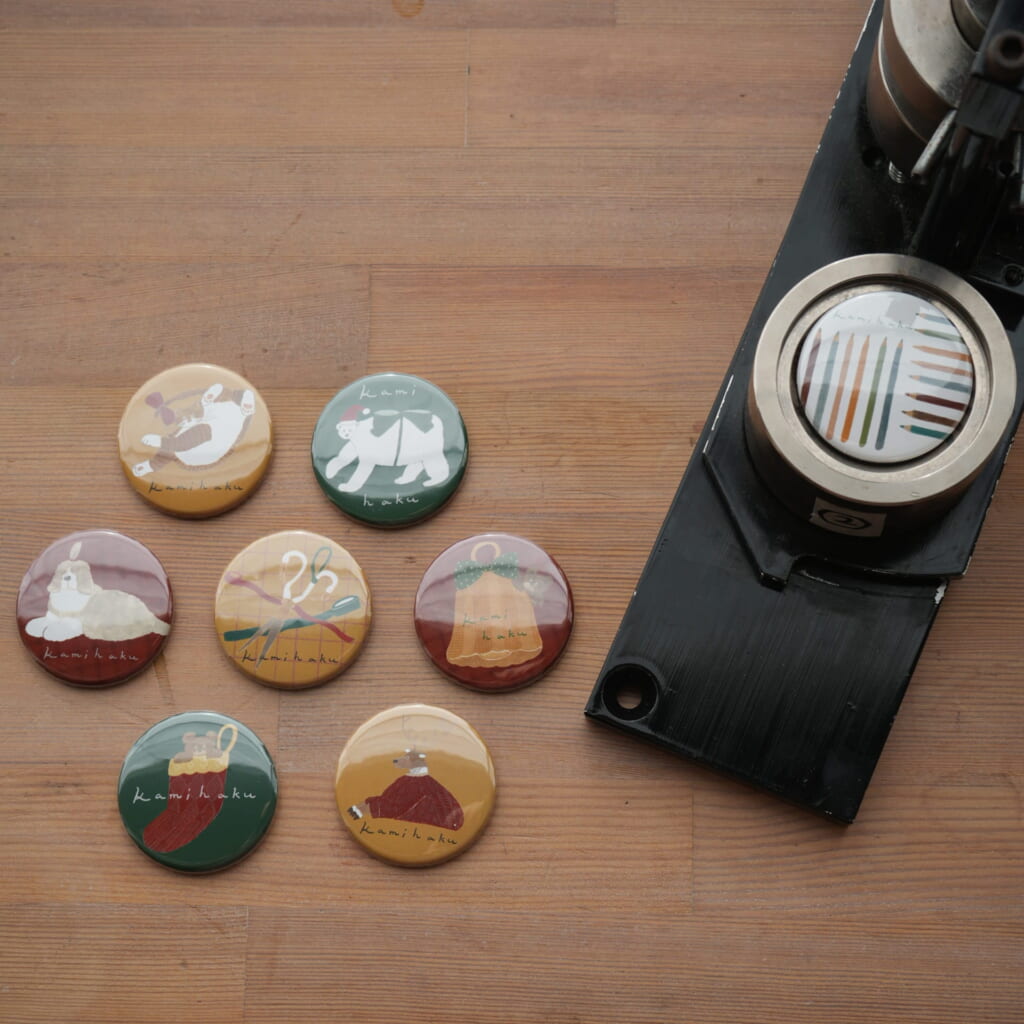 「缶バッジ（全8種）」好きな絵柄を選んだら、専用のマシーンを使ってご自身で缶バッジを作ることができます。（300円／Illustration：トビマツショウイチロウ）